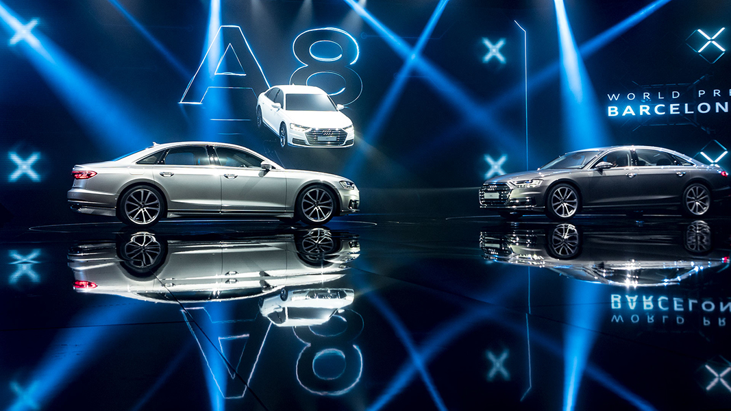 Twei Audi A8 in einer Eventhalle, die sich im glänzenden Boden spiegeln. Sie werden von blauen Scheinwerfern angestrahlt