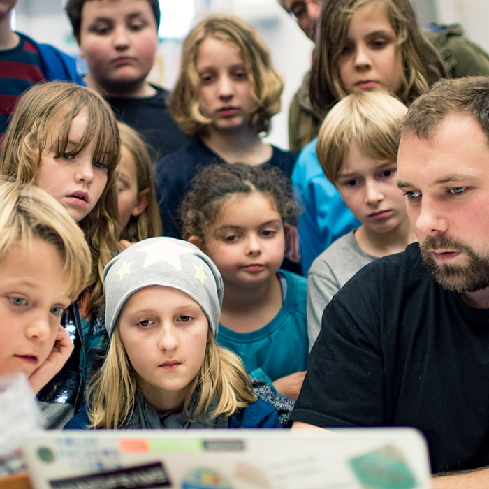 11 Kinder stehen in einem Pulk mit großen Augen hinter und neben CodeWeek-Inspirer Boris Crismancich, der auf seinem Laptop hackt.