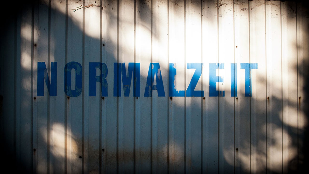 Alter Bauwagen mir grauer Wand, auf der in blauen Blockbuchstaben "NORMALZEIT" steht.