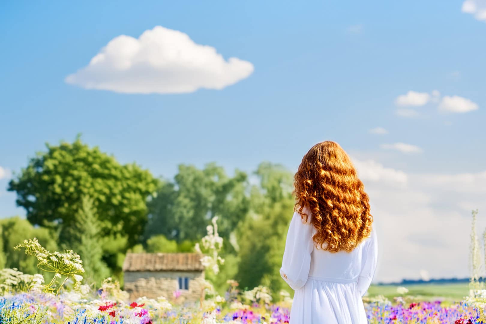 Eine Frau mit roten Haaren steht im weißen Kleid auf einer blühenden Wiese und schaut in Richtung eines alten Häuschens am Waldrand